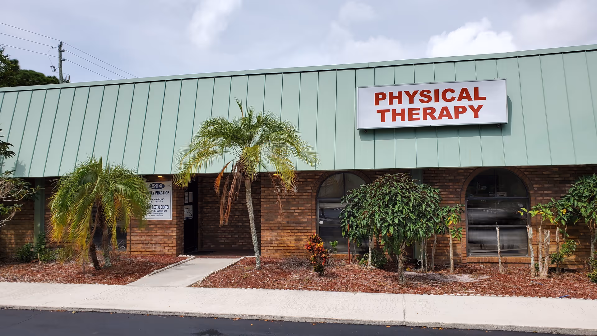 Brick building with green metal awning, palm trees, and a Physical Therapy sign.