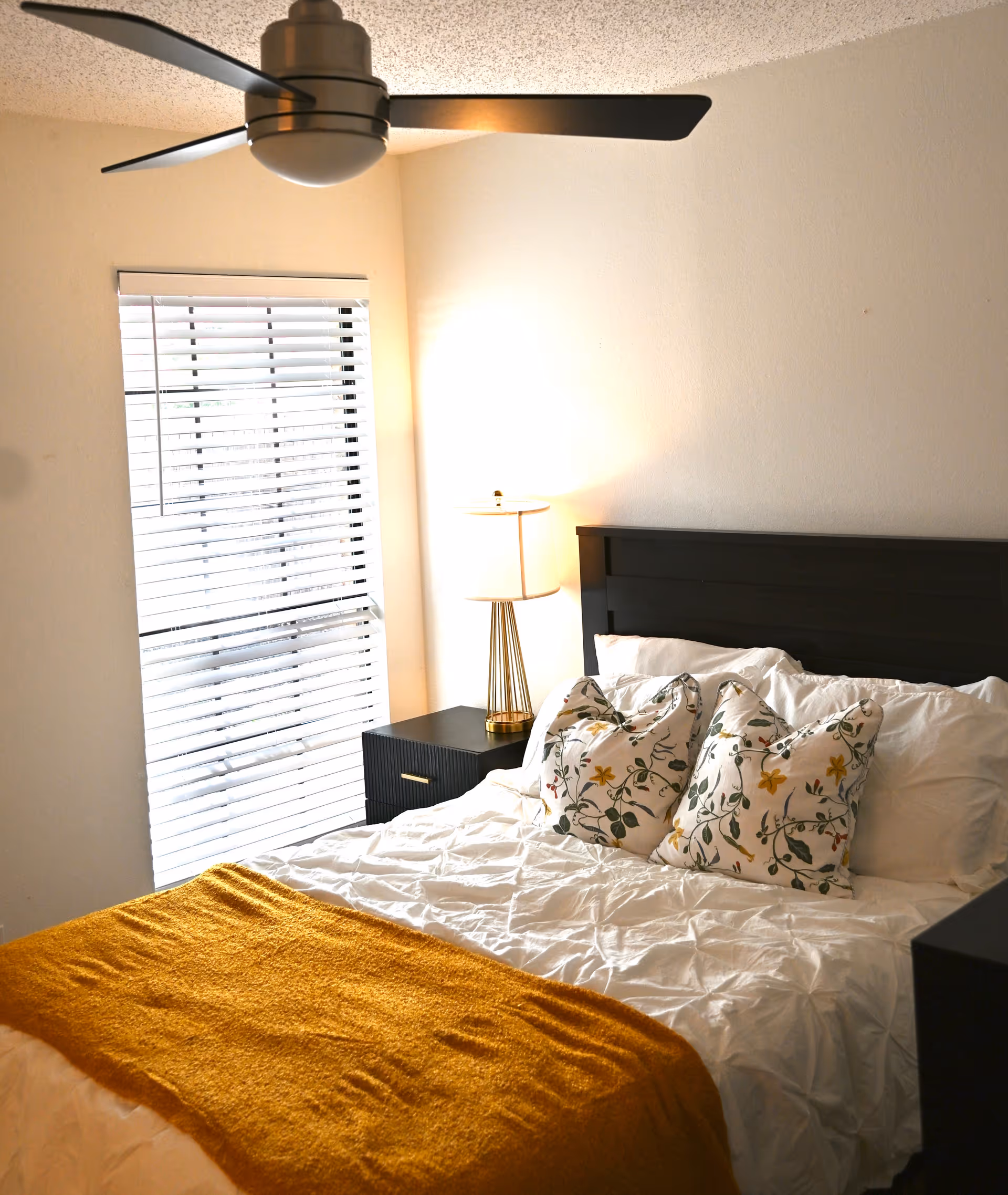 A neatly made bedroom with bed, nightstand, lamp, blinds, and ceiling fan.
