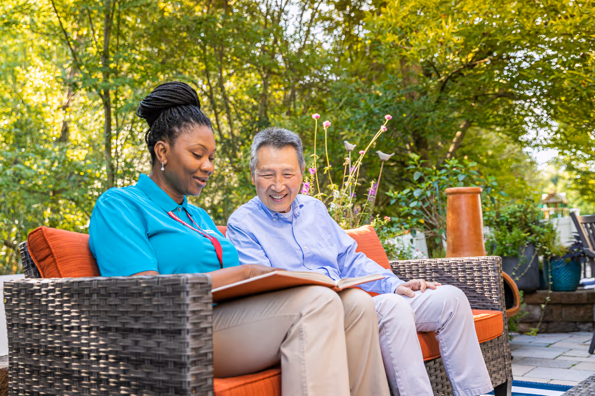 Two people sit outdoors on a wicker couch, smiling while reading a binder.