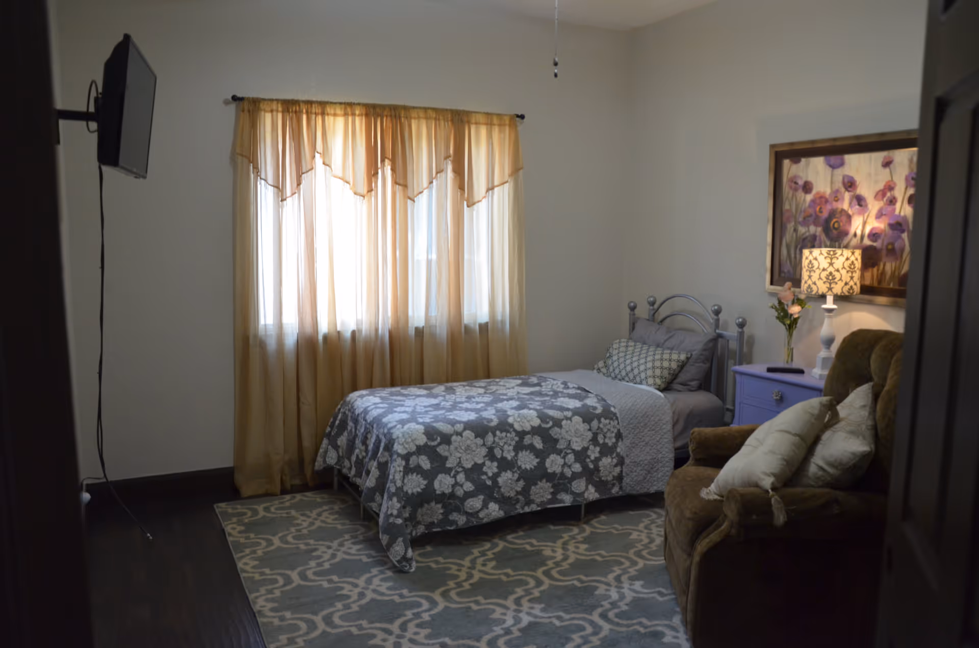 A cozy bedroom with a single bed covered in a floral patterned blanket, a purple nightstand with a lamp and flowers, a comfortable armchair with pillows, a wall-mounted TV, and a window with sheer beige curtains letting in natural light.