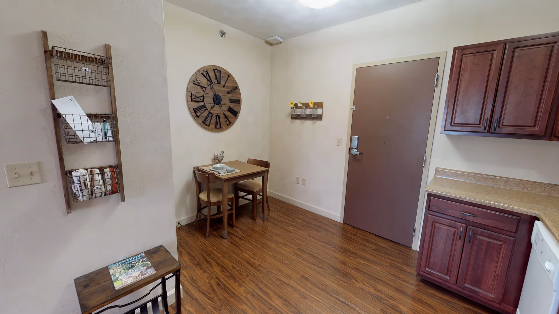 Interior view of a senior living apartment showing a small dining area with a wooden table and two chairs, a large wall clock, a wall-mounted rack with mail and magazines, a door, and kitchen cabinets with a countertop and dishwasher.