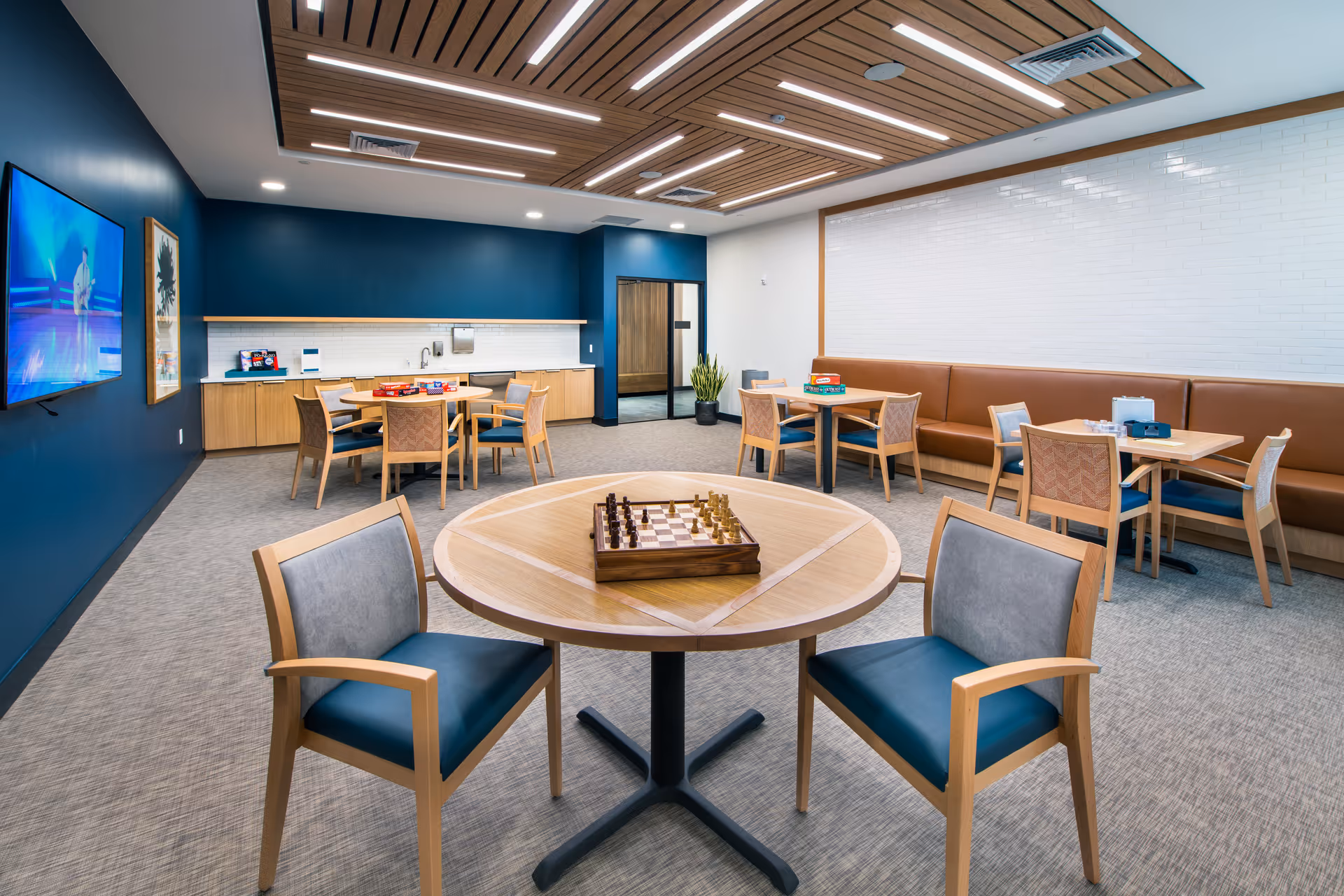 A well-lit common room with multiple wooden tables and chairs arranged for games and social activities. A chessboard is set up on the round table in the foreground. The room features a blue accent wall with a mounted TV, a white tiled wall with a long brown cushioned bench, and a wooden ceiling with recessed lighting. There are board games on some tables and a small kitchenette area with cabinets and a sink in the background.