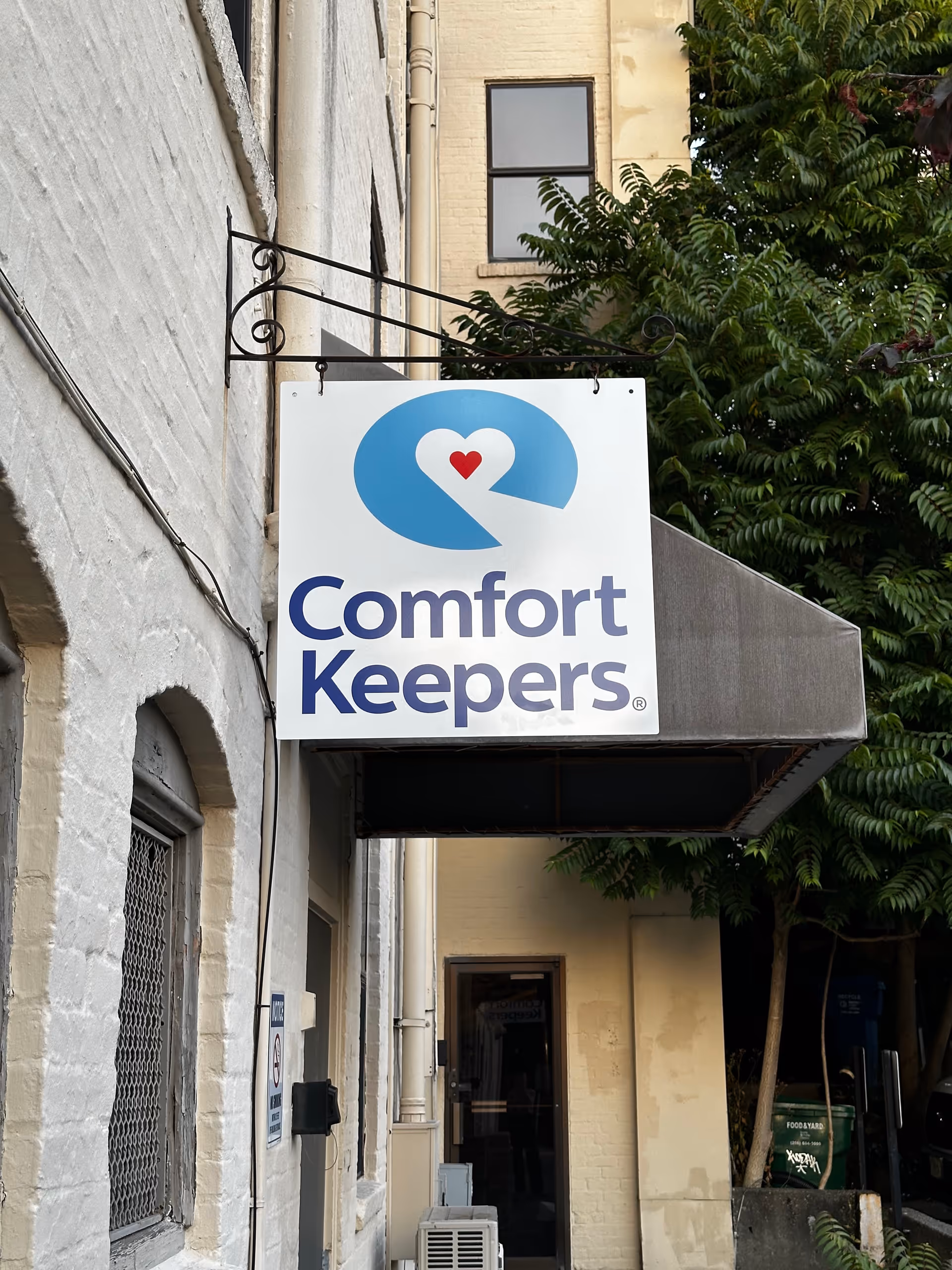 A Comfort Keepers sign hangs outside a beige brick building.