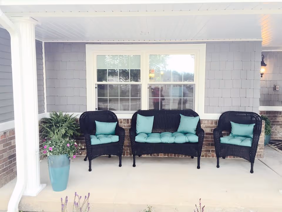 Covered outdoor porch area with three black wicker seats, including a loveseat and two armchairs, all with light blue cushions. There is a large window with white trim behind the seating and two large potted plants with greenery and flowers on either side of the seating area. The porch has a light-colored concrete floor and gray siding with brick accents on the lower wall.
