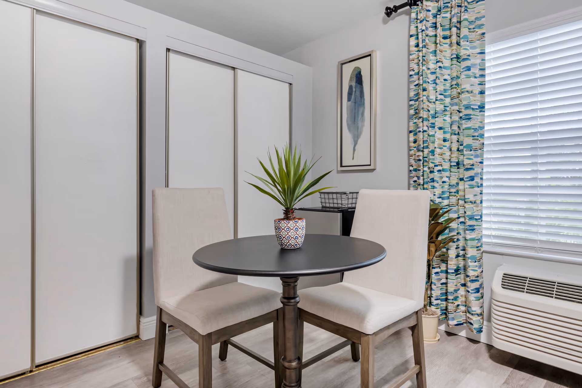 A small dining area with round table, two beige upholstered chairs, potted plant, and window blinds.