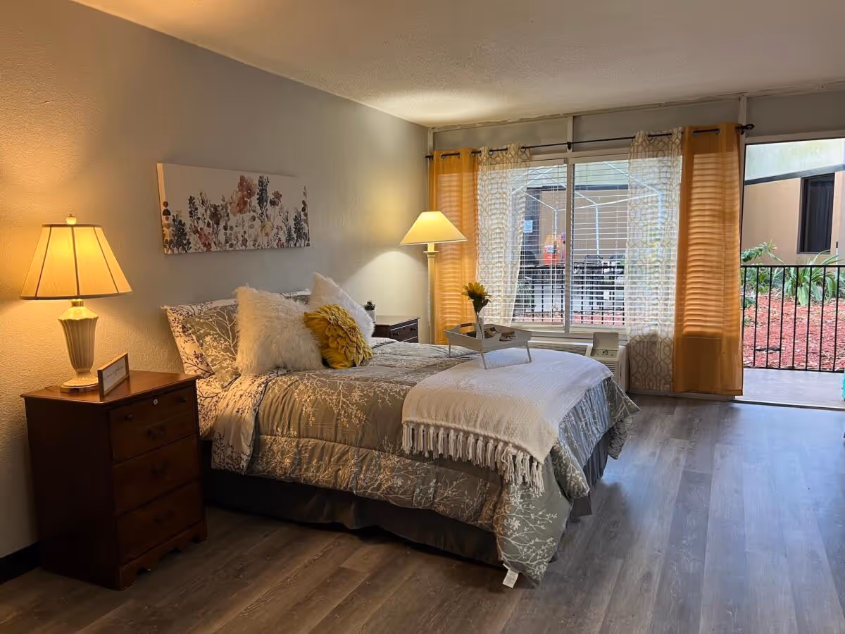 A furnished bedroom with a made bed, bedside tables and lamps, and a large window/door opening to an outdoor patio.