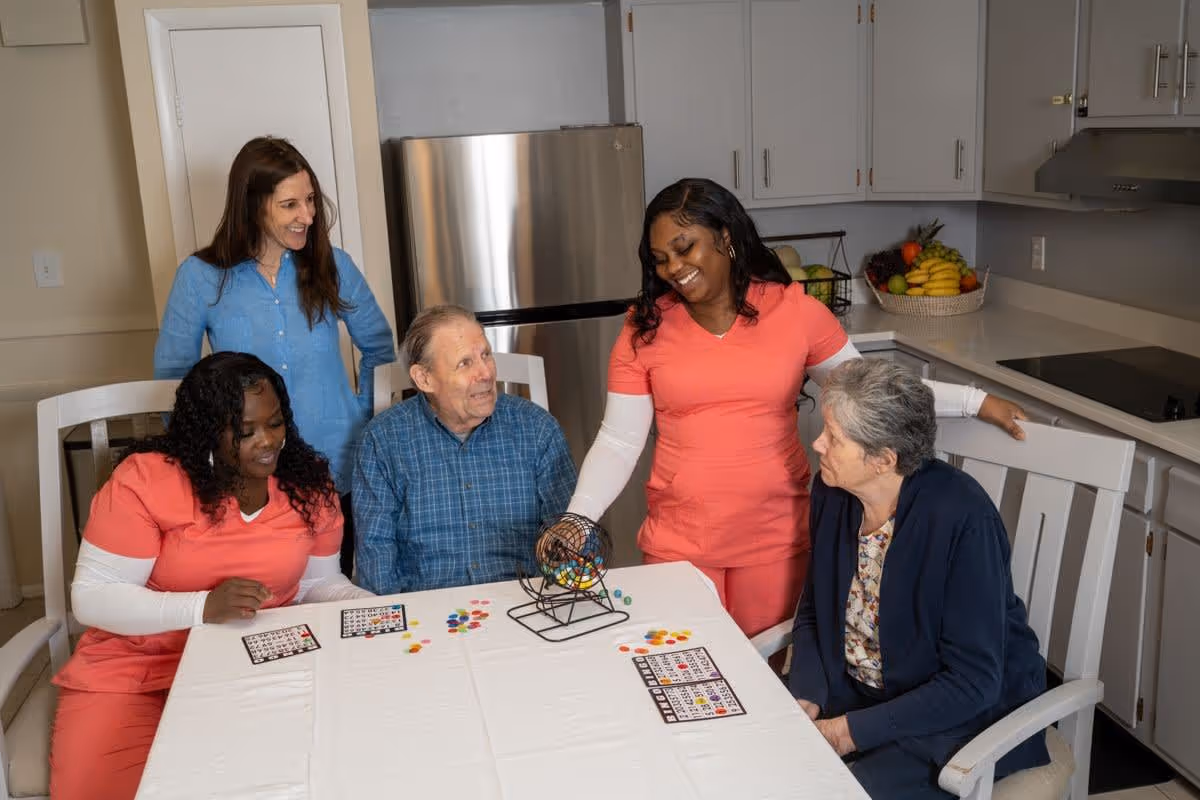 Two elderly individuals and three caregivers gathered around a table in a kitchen playing a game with bingo cards and colorful markers. The caregivers are smiling and engaging with the elderly participants, creating a warm and friendly atmosphere.