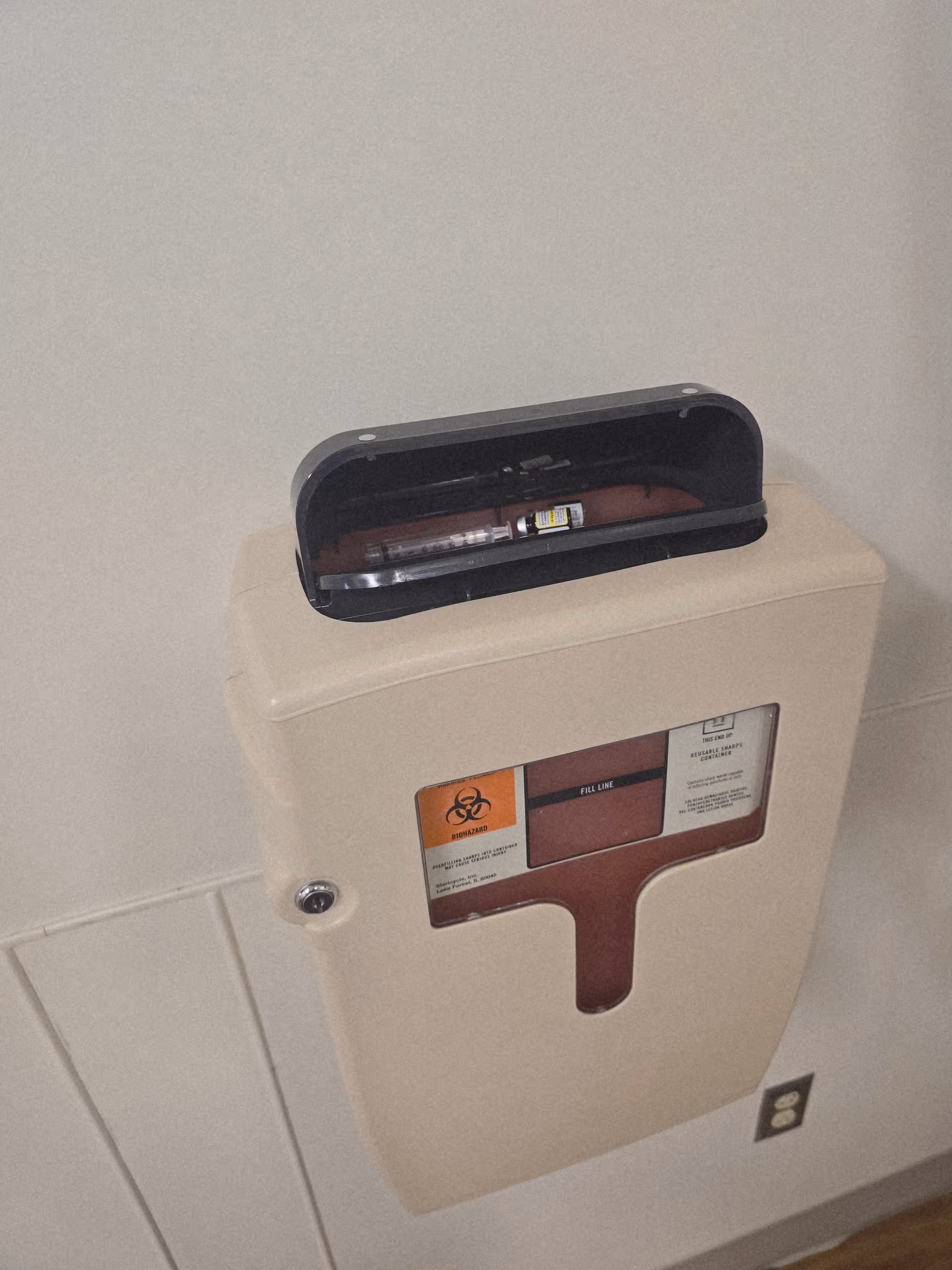 Beige wall mounted sharps disposal container with opened lid and biohazard label.