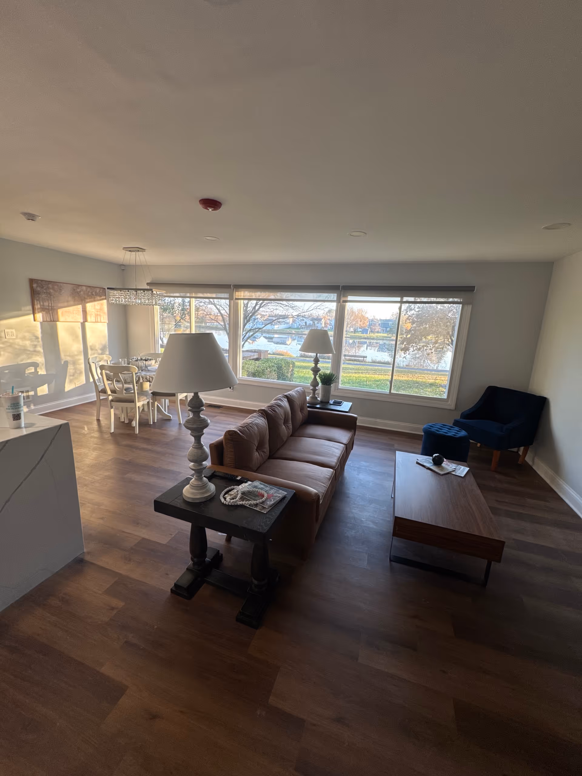 Bright living room with brown sofa, coffee table, lamps, and large windows overlooking water.