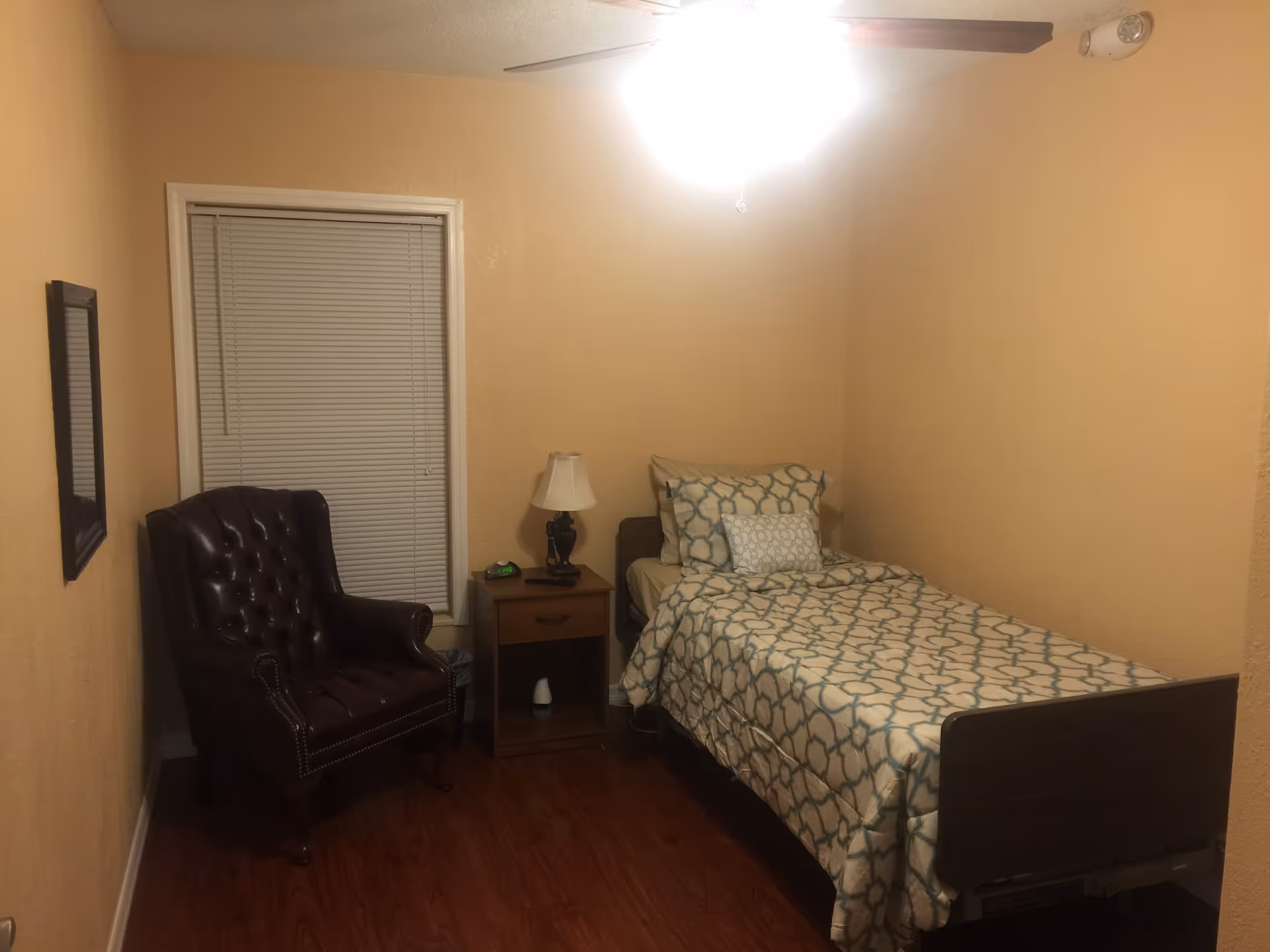 Small bedroom with a twin bed in patterned bedding, a wooden nightstand with a lamp, a leather armchair, and a window with closed blinds.