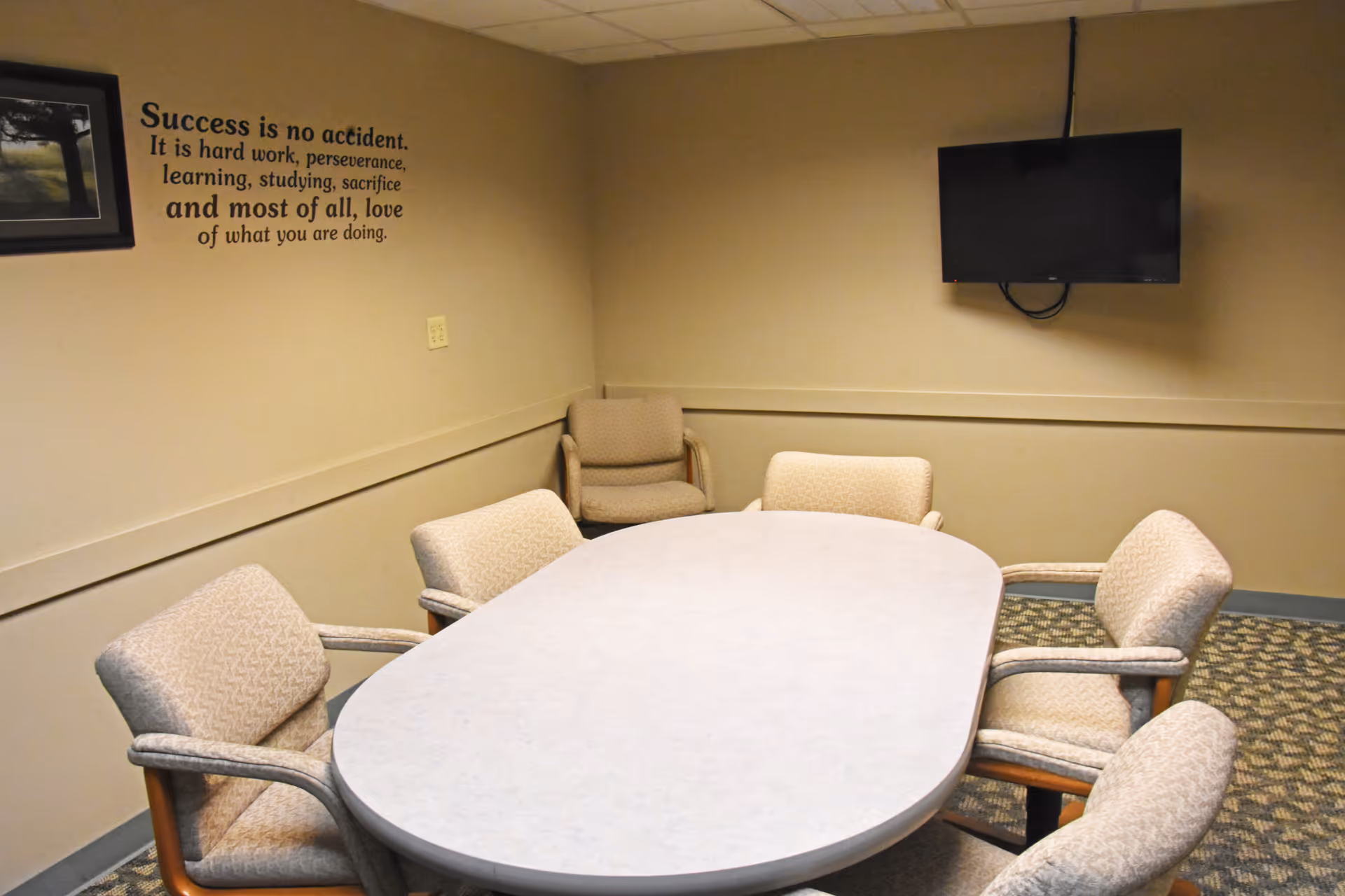 An interior meeting room with a large oval table, chairs, and a wall-mounted TV.