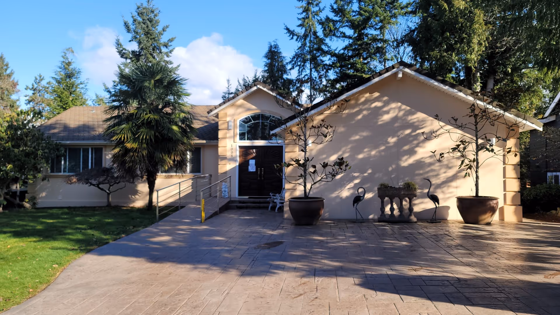 A single-story beige senior living facility entrance with potted trees and a paved front walkway.
