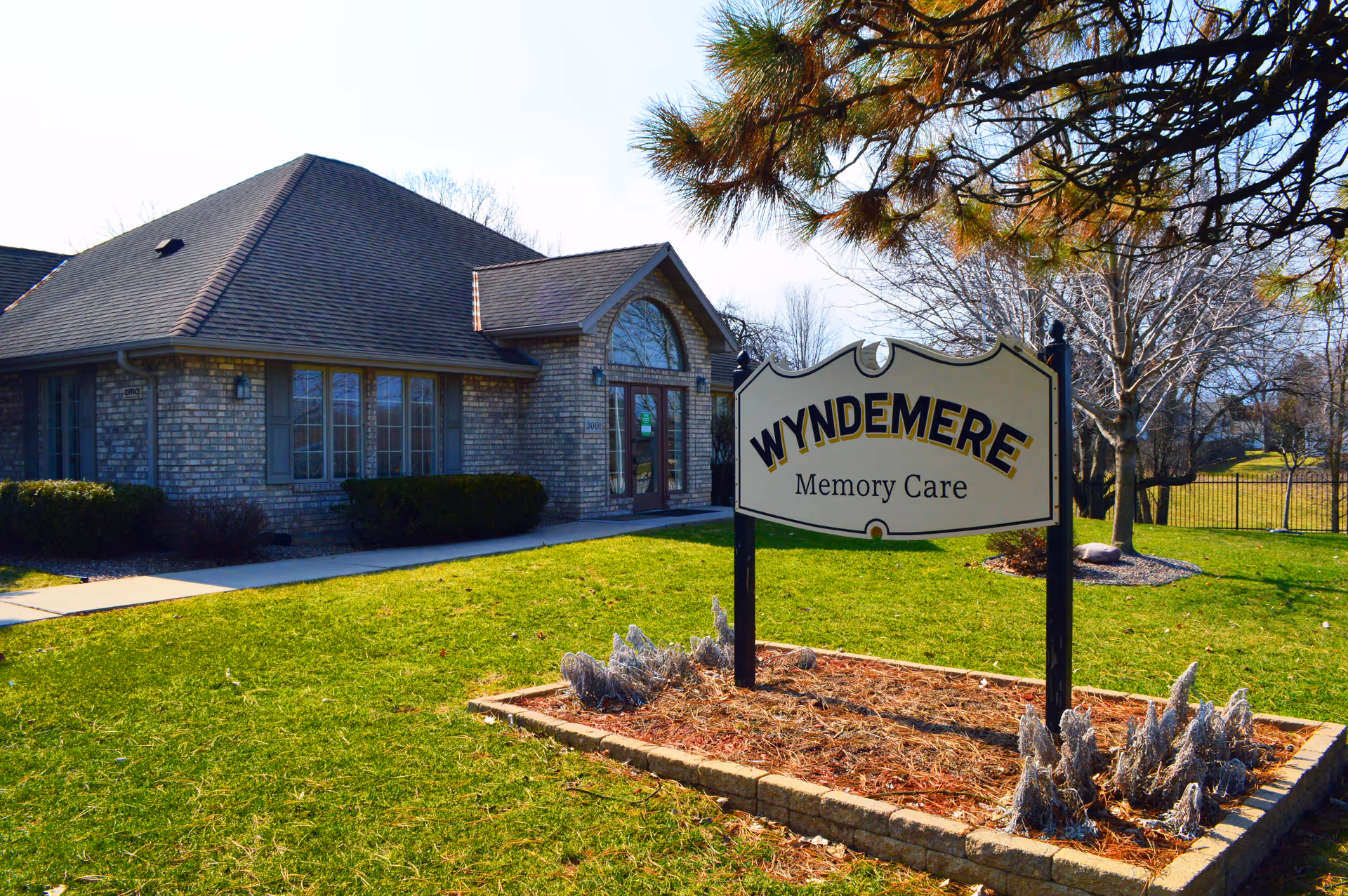 Exterior view of Wyndemere Memory Care facility showing a brick building with a dark shingled roof, large windows, and a glass entrance door. In front of the building is a sign that reads 'WYNDEMERE Memory Care' placed in a landscaped area with grass, small plants, and trees in the background.