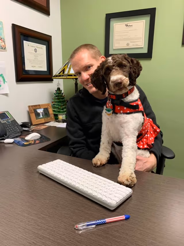 A man sits at a desk with a spotted dog wearing a red scarf.