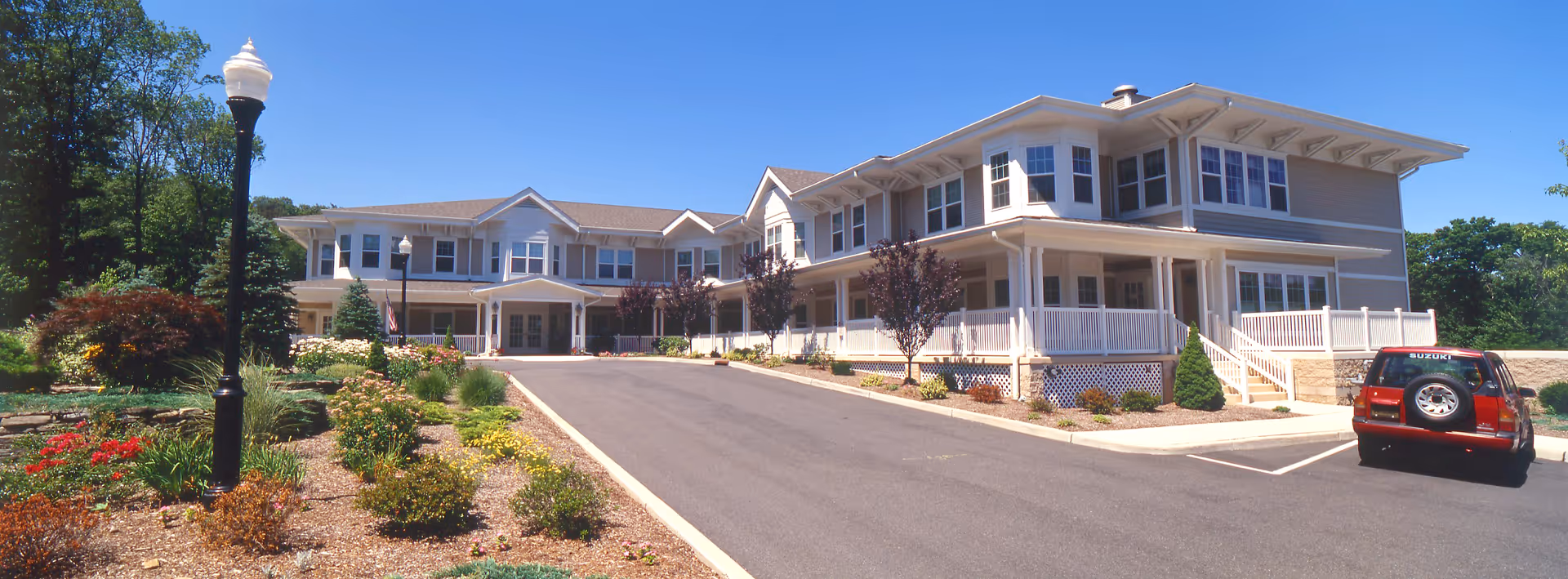 Exterior view of a large two-story senior living facility building with beige siding and white trim, surrounded by landscaped gardens with flowers and shrubs, a paved driveway, a street lamp, and a red Suzuki SUV parked in a parking area under a clear blue sky.