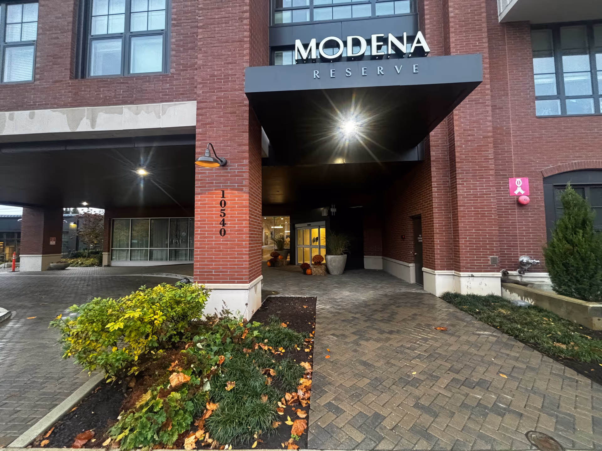 Entrance to Modena Reserve senior living facility with a covered driveway, brick exterior, potted plants, and the address number 10540 displayed on a brick column.