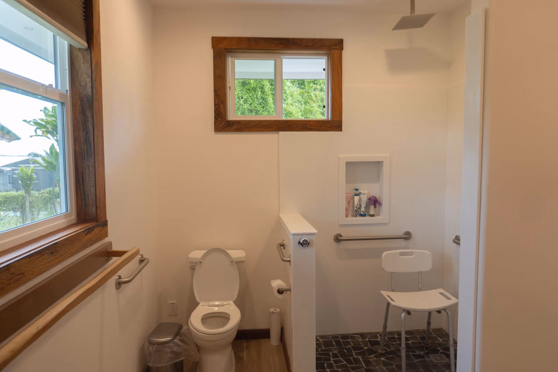 Accessible bathroom with toilet, grab bars, window, and a small chair.