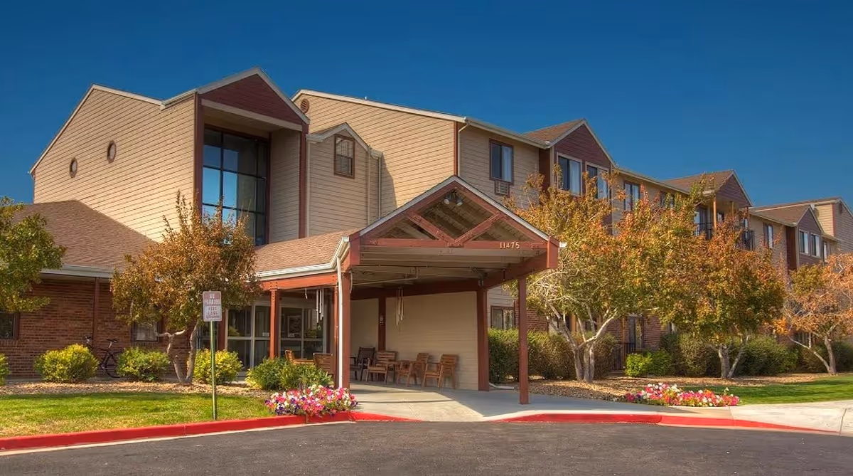 Exterior view of Northglenn Heights Assisted Living and Memory Care building with a covered entrance, several chairs under the entrance canopy, landscaped bushes and trees with autumn foliage, and a clear blue sky.