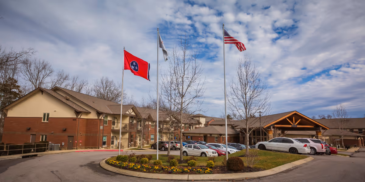 Exterior view of The Lodge at Natchez Trace senior living facility showing a curved driveway with a landscaped island featuring three flagpoles with flags, parked cars, and a large building with a peaked wooden entrance under a partly cloudy sky.