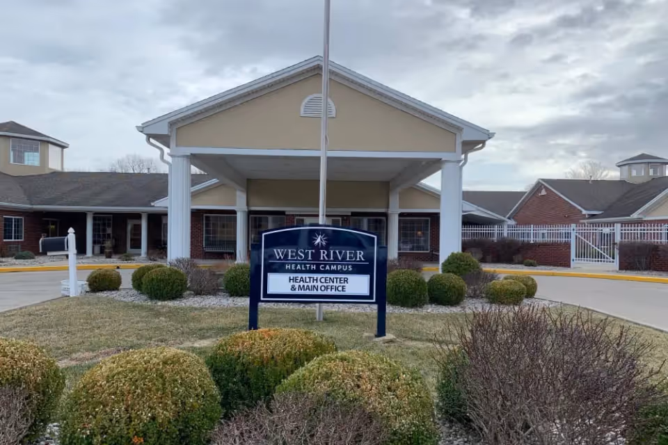 Front entrance of a health campus building with a sign reading 'West River Health Campus Health Center & Main Office' and landscaped bushes in front.