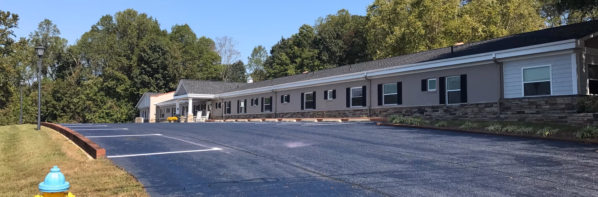 Single-story senior living building with stone veneer, gray siding, parking, and surrounding trees.