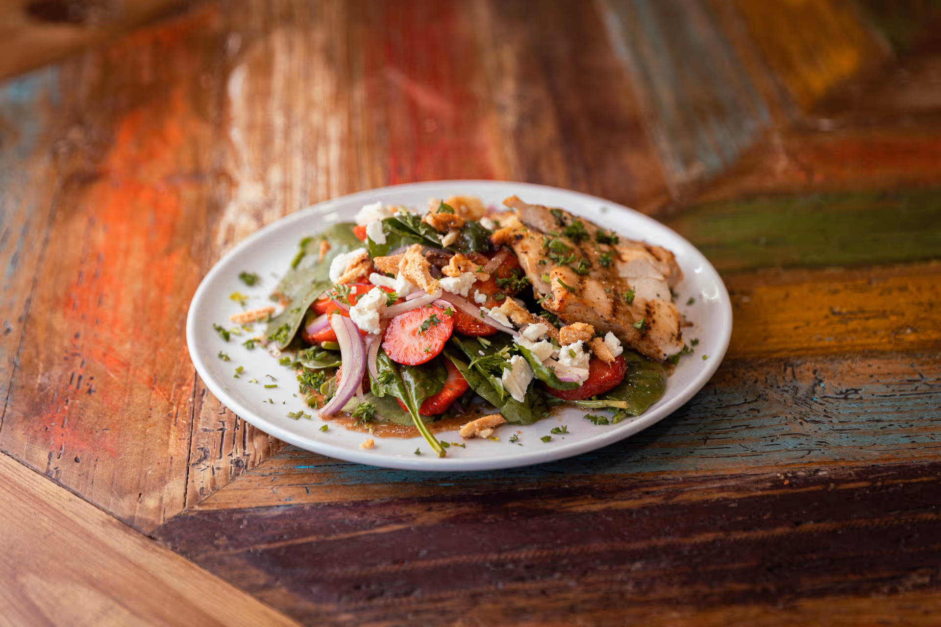 A white plate with a fresh salad consisting of spinach leaves, sliced strawberries, red onion, crumbled cheese, and grilled chicken pieces, placed on a colorful wooden table.