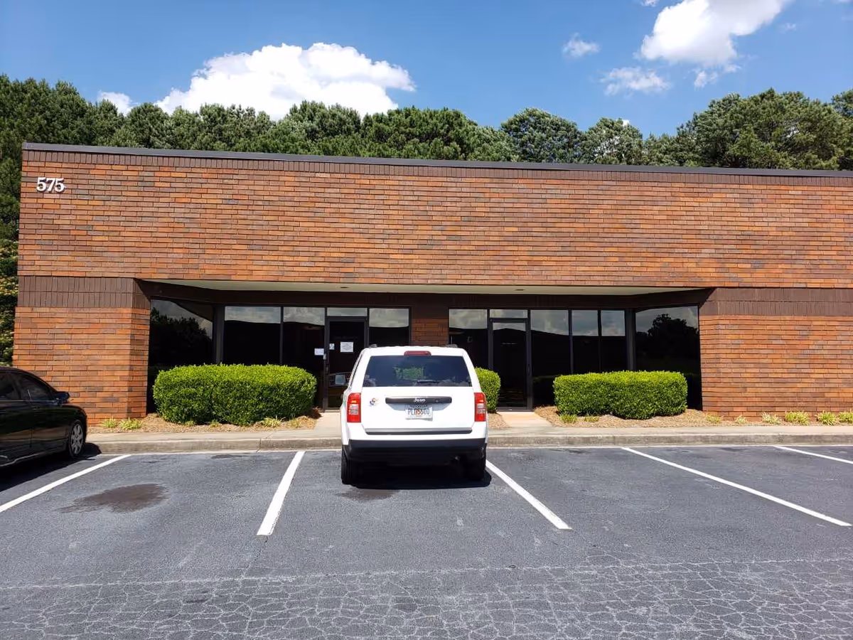 Front view of a brick building with glass entrance, parking, and trimmed hedges.