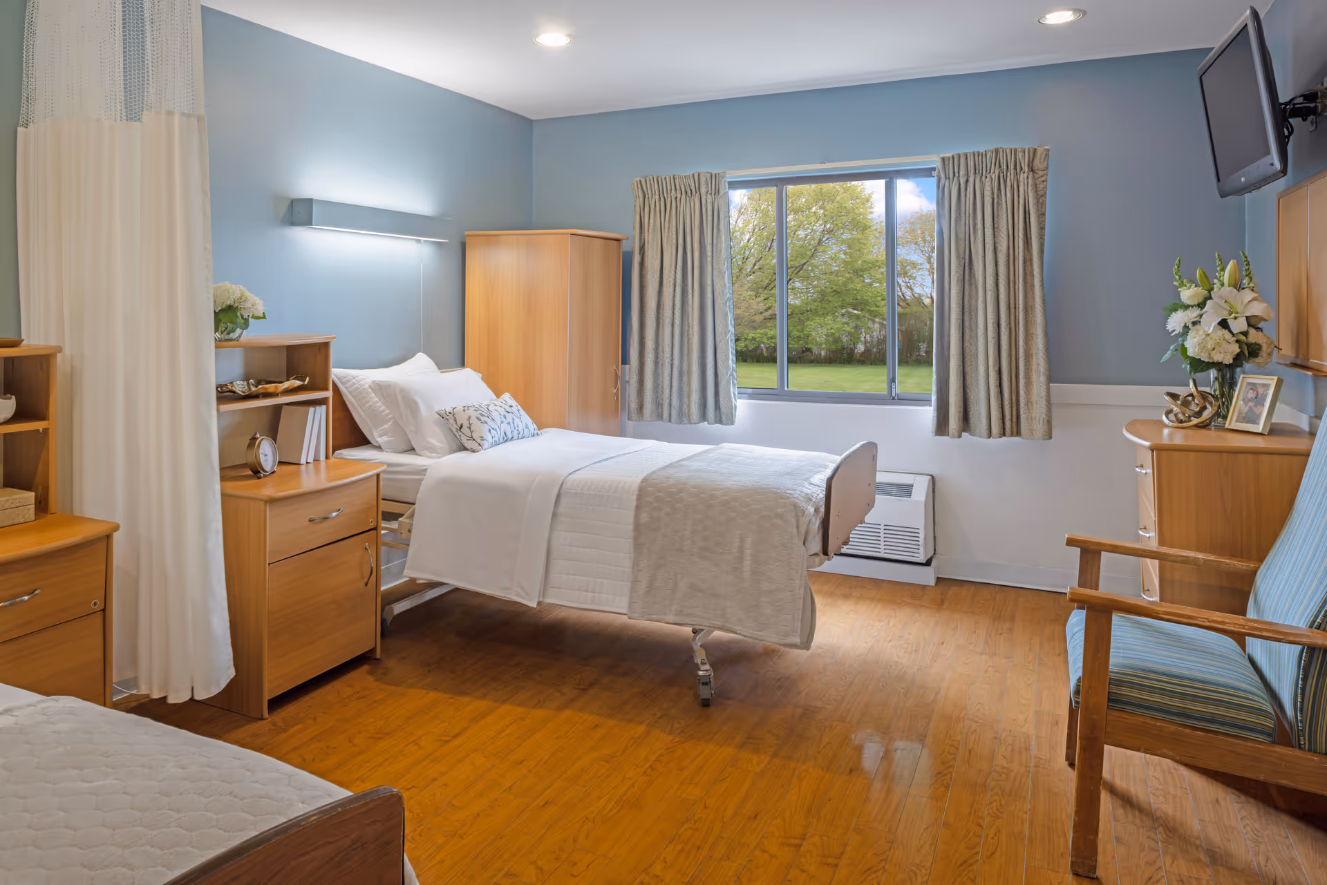 A furnished senior living bedroom in Bella Terra Lombard with bed, dresser, chair, window.