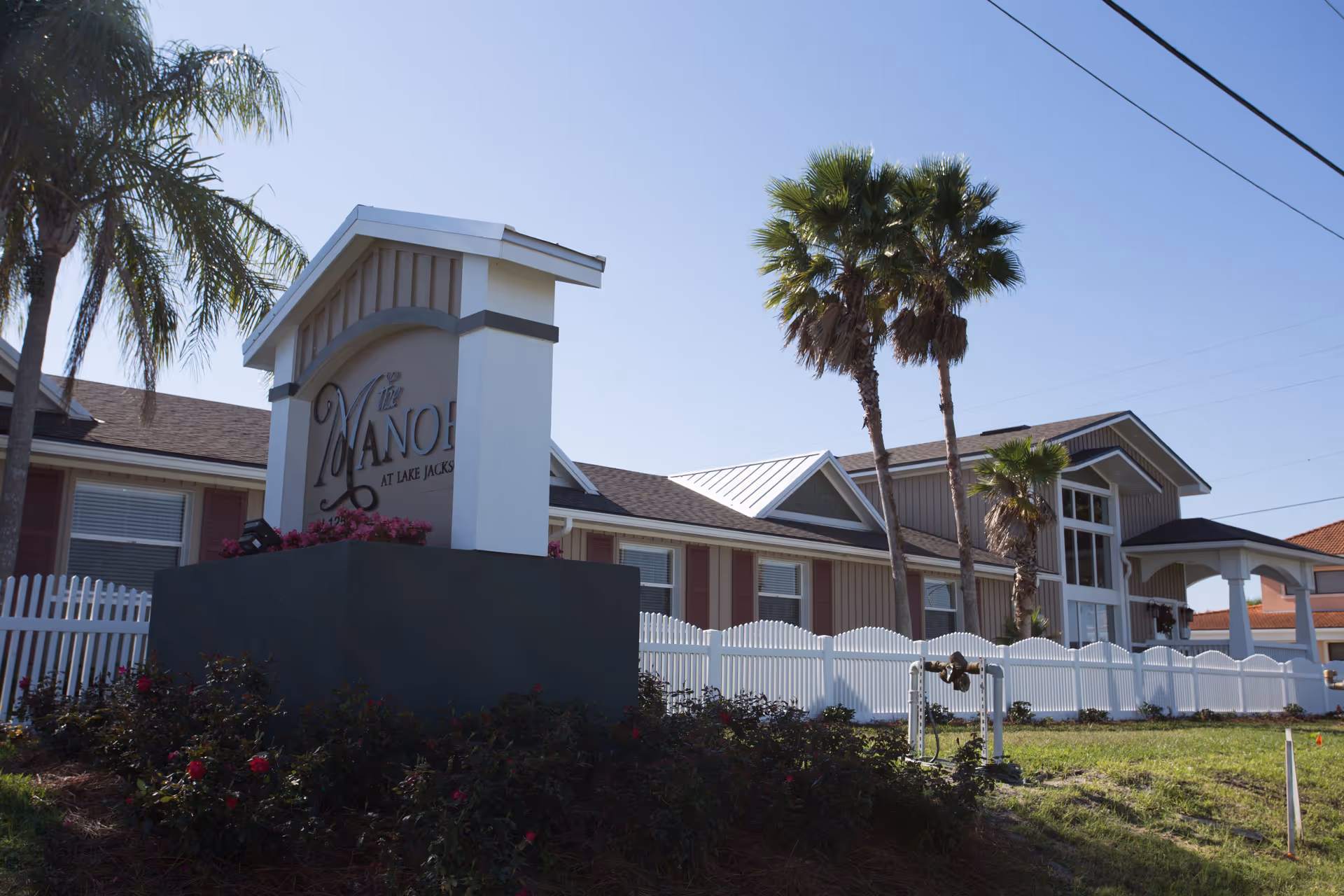 Exterior view of The Manor at Lake Jackson facility with a large sign in front, white picket fence, palm trees, and a clear blue sky.