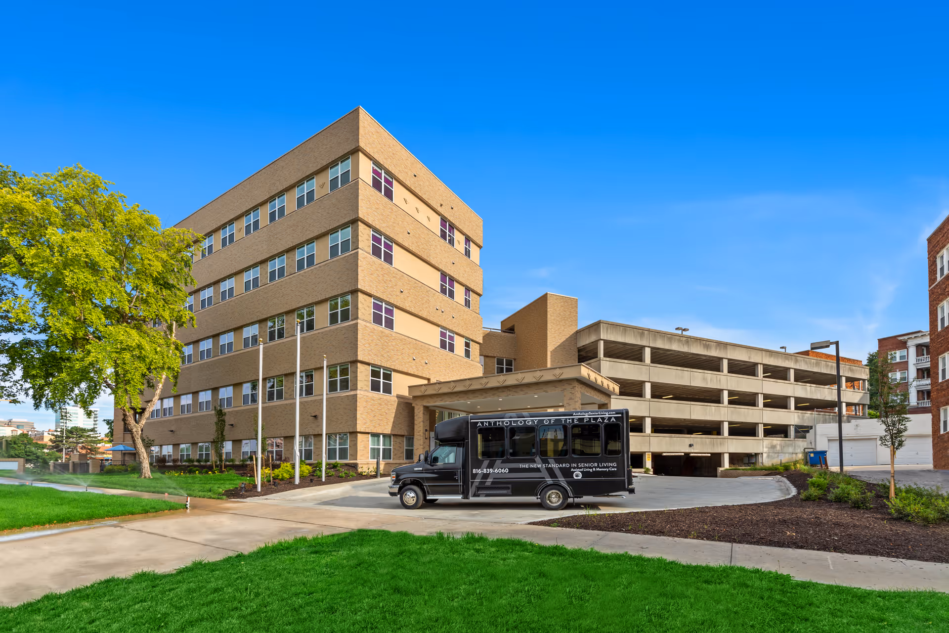 Exterior view of a multi-story senior living facility building with a parking garage attached. A black shuttle bus labeled 'Anthology of the Plaza' is parked in front of the building. The sky is clear and blue, and there is green grass and a tree on the left side of the image.