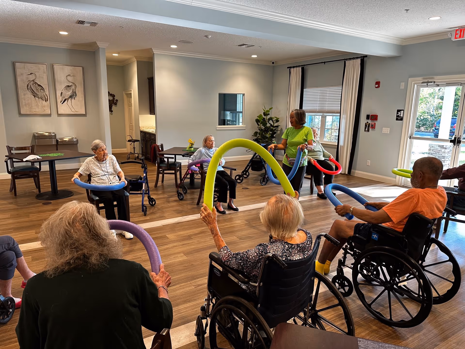 A group of elderly individuals, some in wheelchairs, participate in a seated exercise class using colorful foam noodles in a spacious, well-lit room with wooden floors and large windows. A woman in a green shirt leads the activity while others follow along.