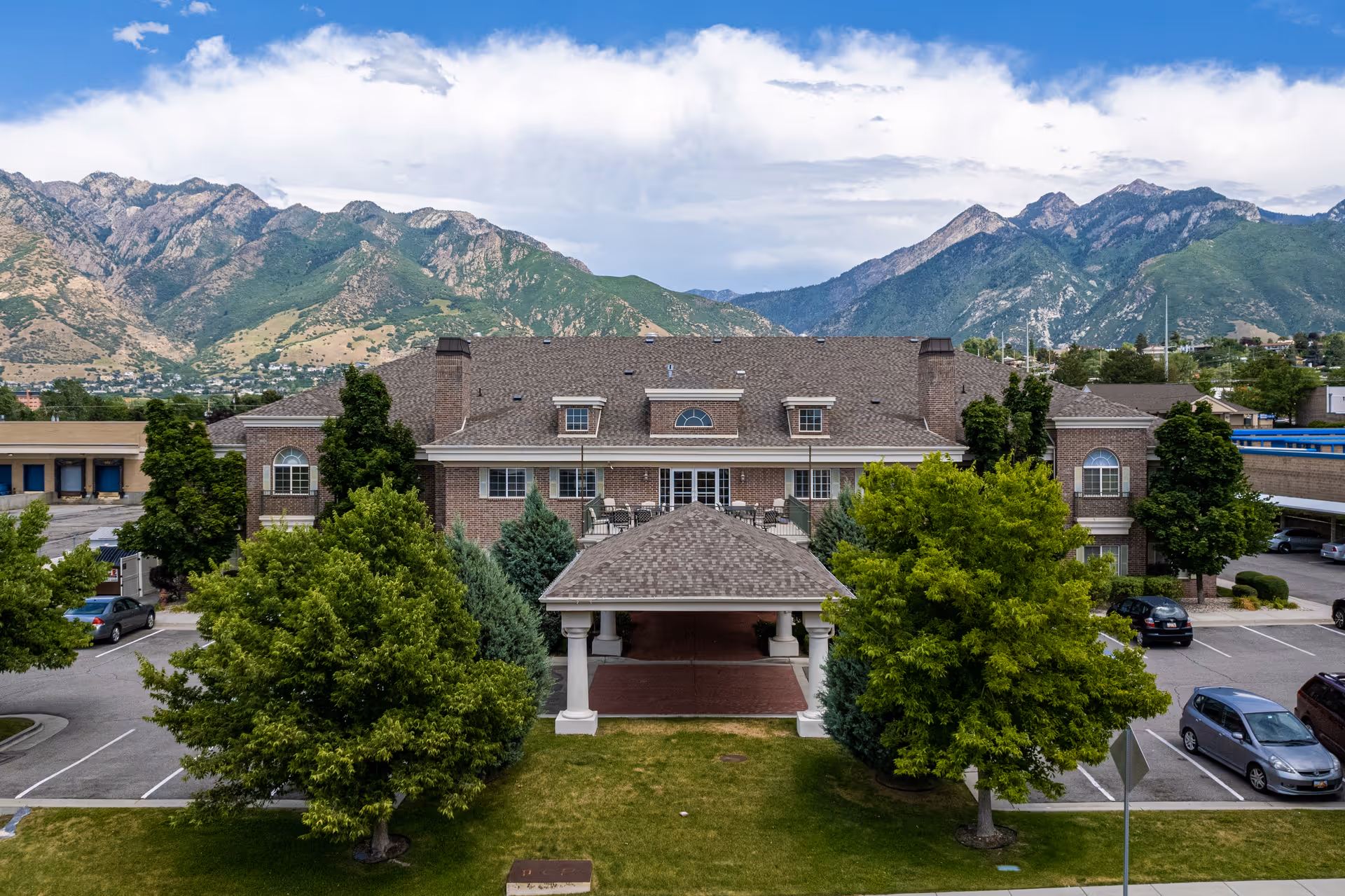 A brick senior living building with a covered entrance and surrounding parking lot against mountains.