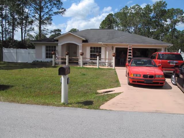 A single-story building with a front entrance, driveway, mailbox, and red cars in the driveway.