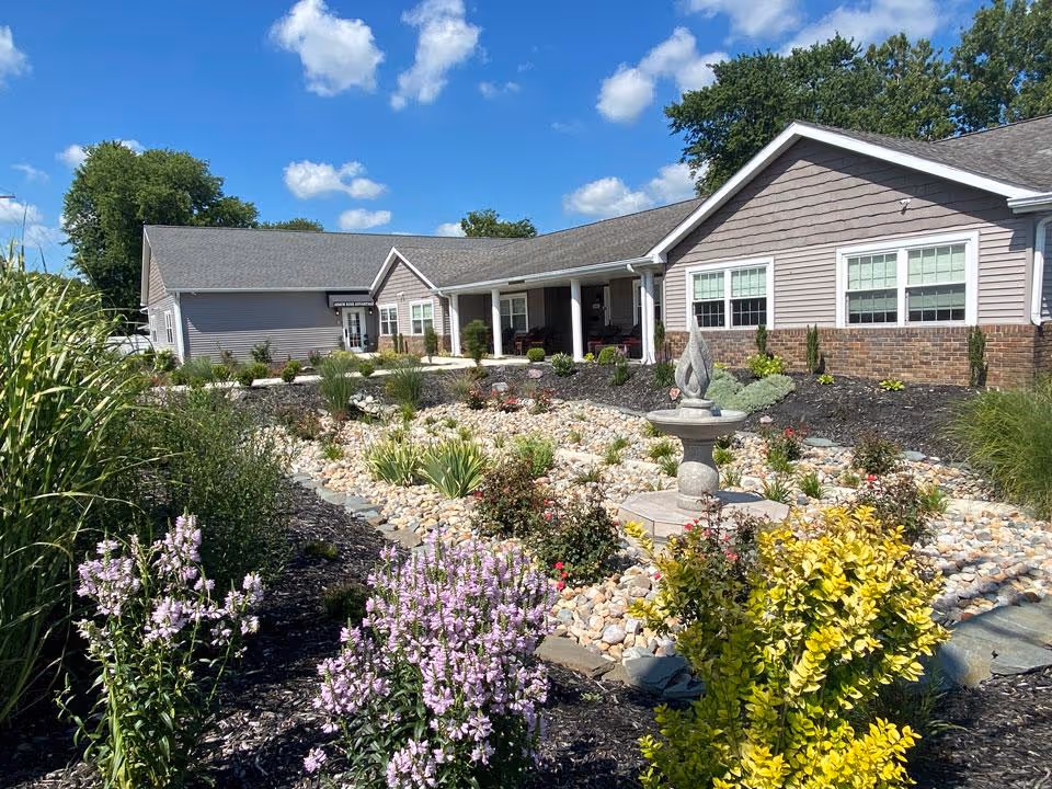 Front exterior of a single-story senior living building with landscaped rock beds, flowering shrubs, and a stone fountain under a blue sky.