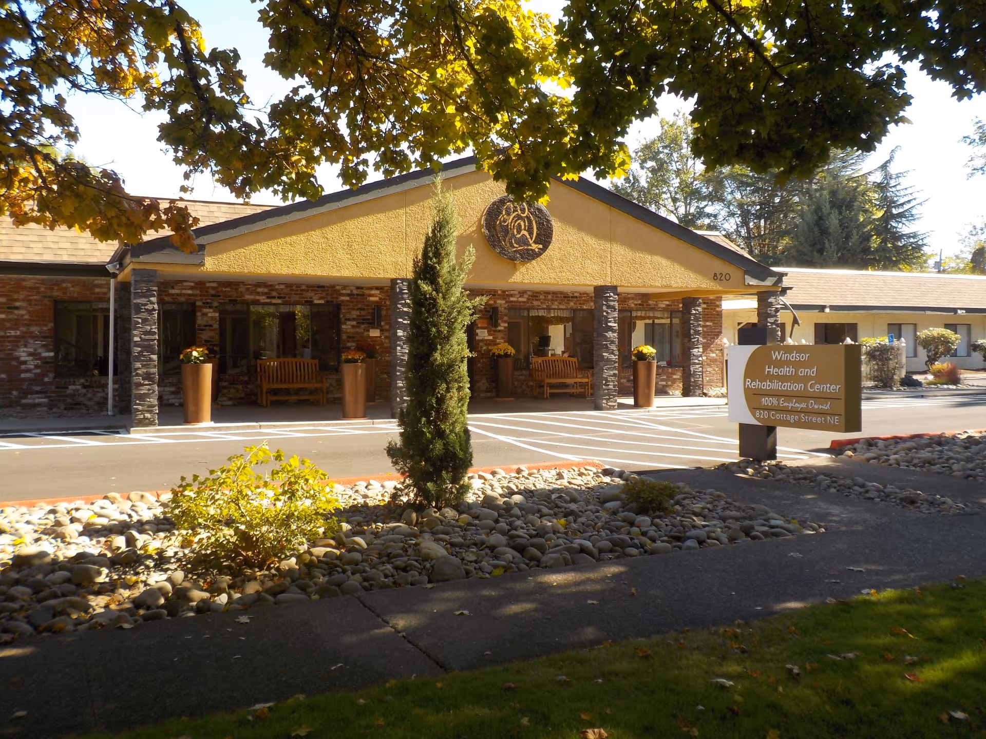 Front entrance of the Windsor Health and Rehabilitation Center building with a sign, covered entry, benches, and landscaped rock beds.