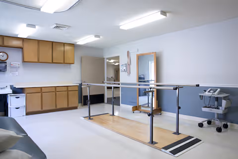 Physical therapy room with parallel walking bars, a wall mirror, cabinets and medical equipment.