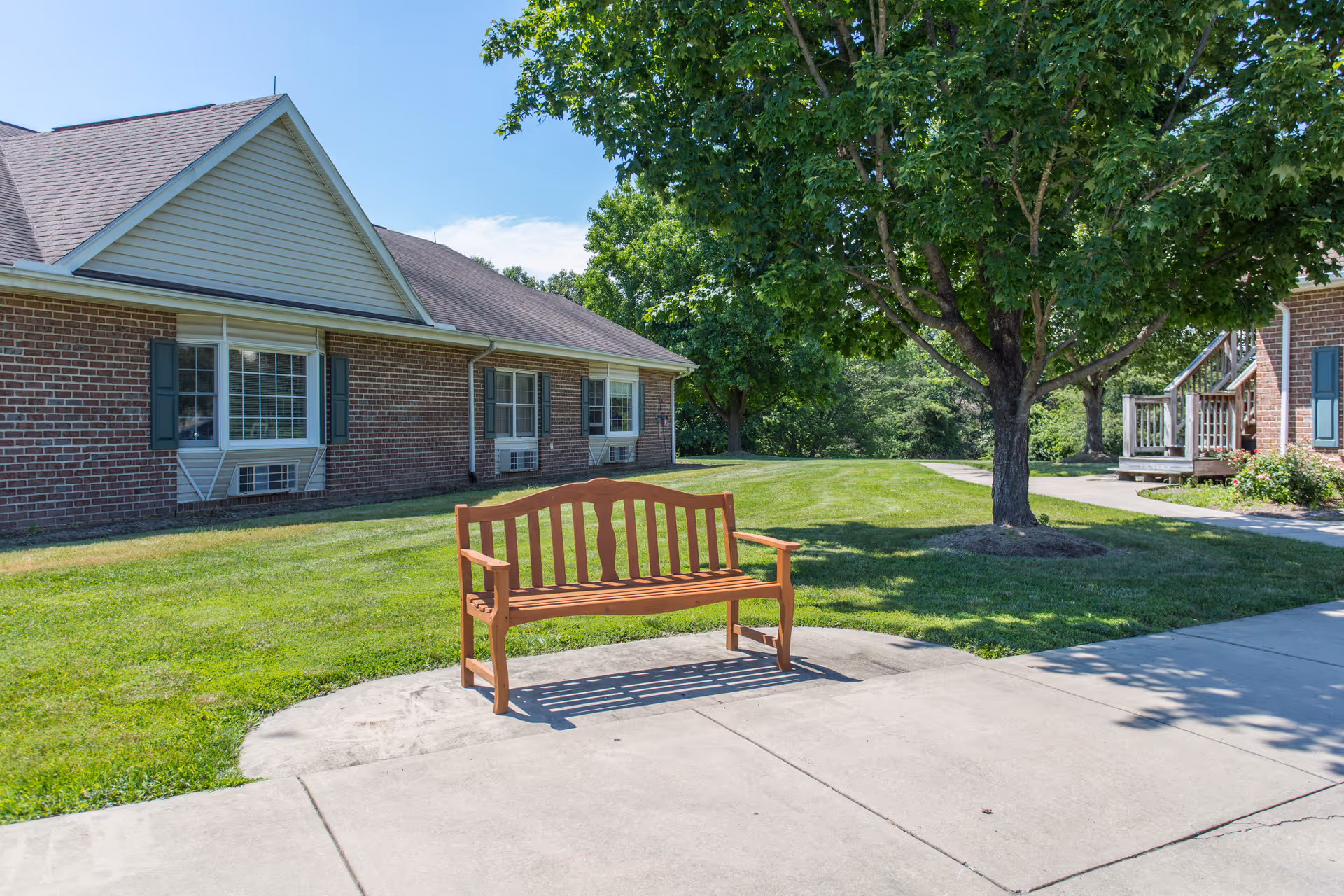 A wooden bench on a concrete path in front of a brick senior living building with a lawn and tree.