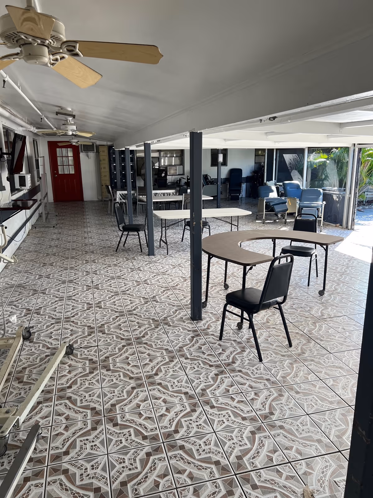 A spacious room with patterned tile flooring, several tables and chairs arranged throughout, ceiling fans, and large windows letting in natural light. The room has a red door at the far end and some reclining chairs near the windows.