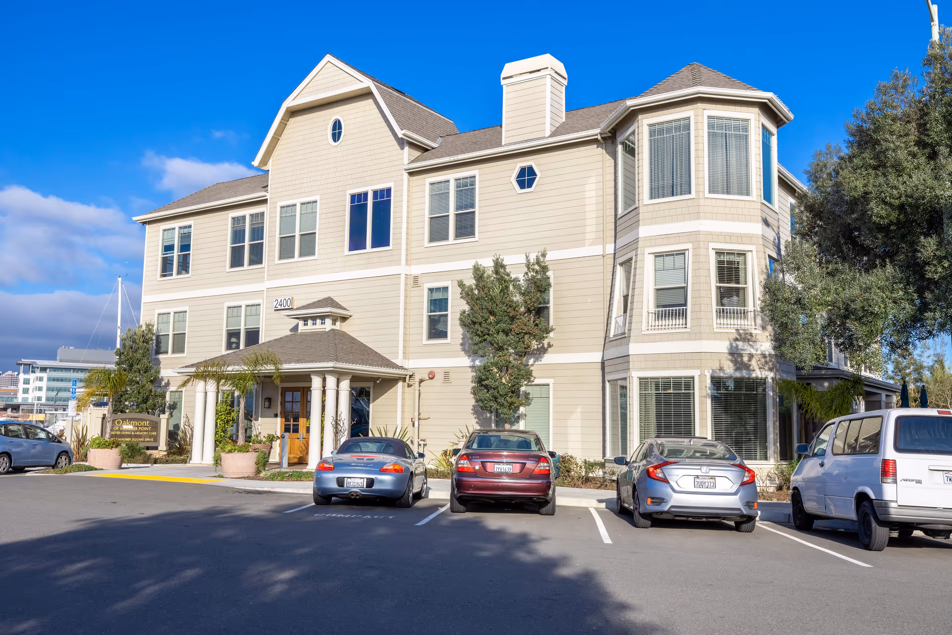 Exterior view of a three-story senior living facility building named Oakmont of Mariner Point with several parked cars in front under a clear blue sky.