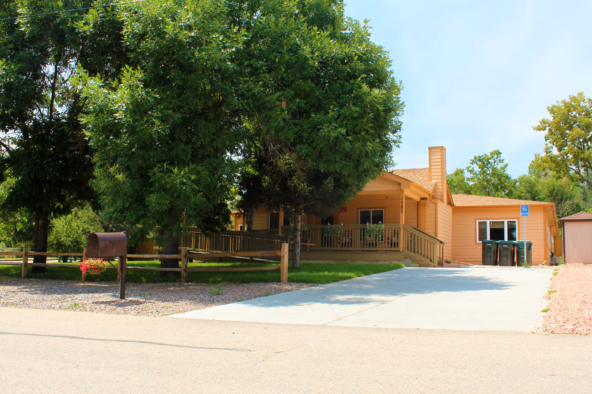 Exterior view of a single-story building with a beige facade, a wooden ramp leading to the entrance, surrounded by green trees and a small fenced area with a mailbox and flowers. There is a concrete driveway with a handicapped parking sign and trash bins near the building.