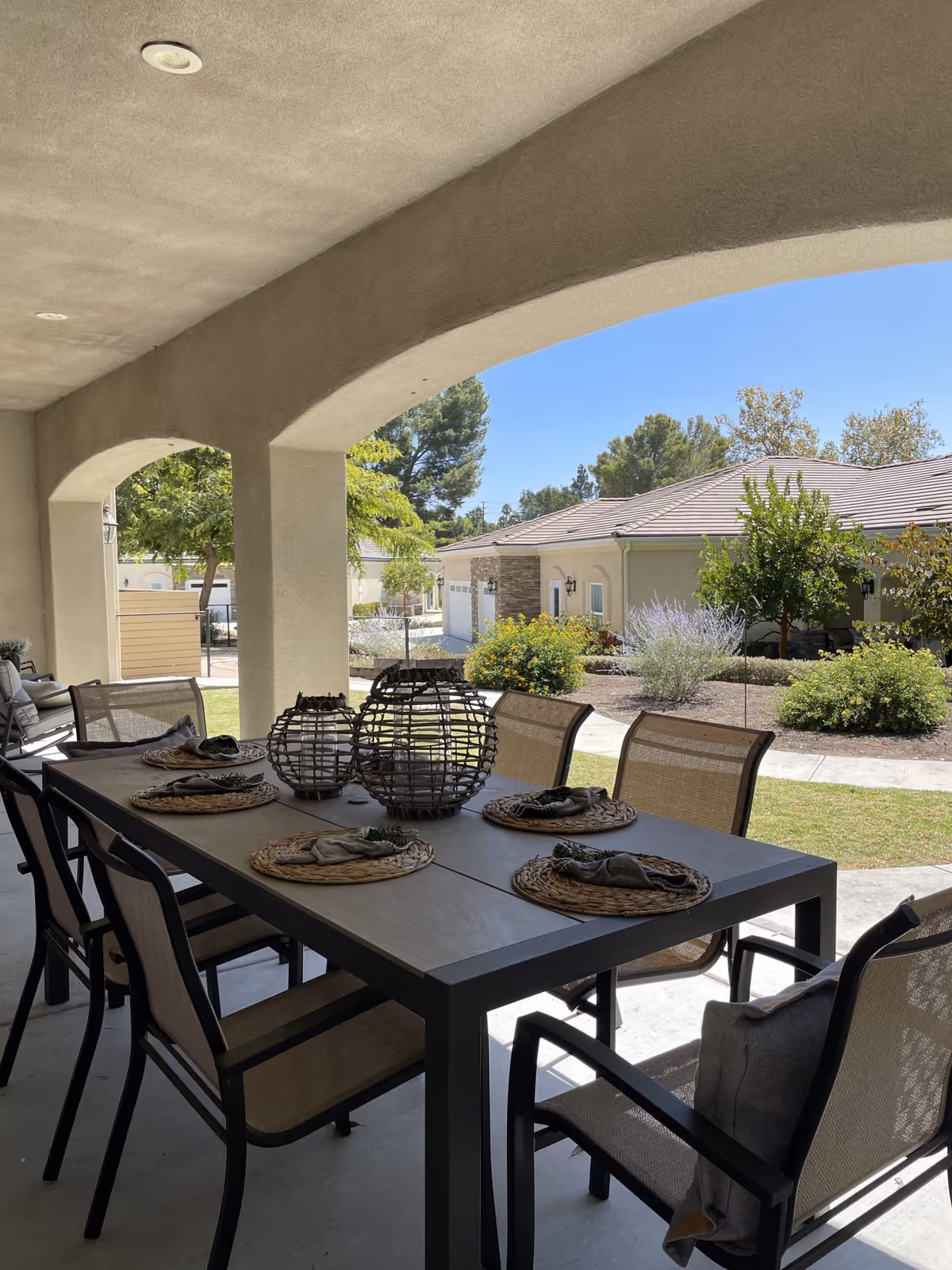 Covered outdoor patio area with a large rectangular dining table set with six chairs. The table has decorative placemats, napkins, and three spherical lanterns as centerpieces. In the background, there is a view of a garden with bushes, trees, and a neighboring building under a clear blue sky.