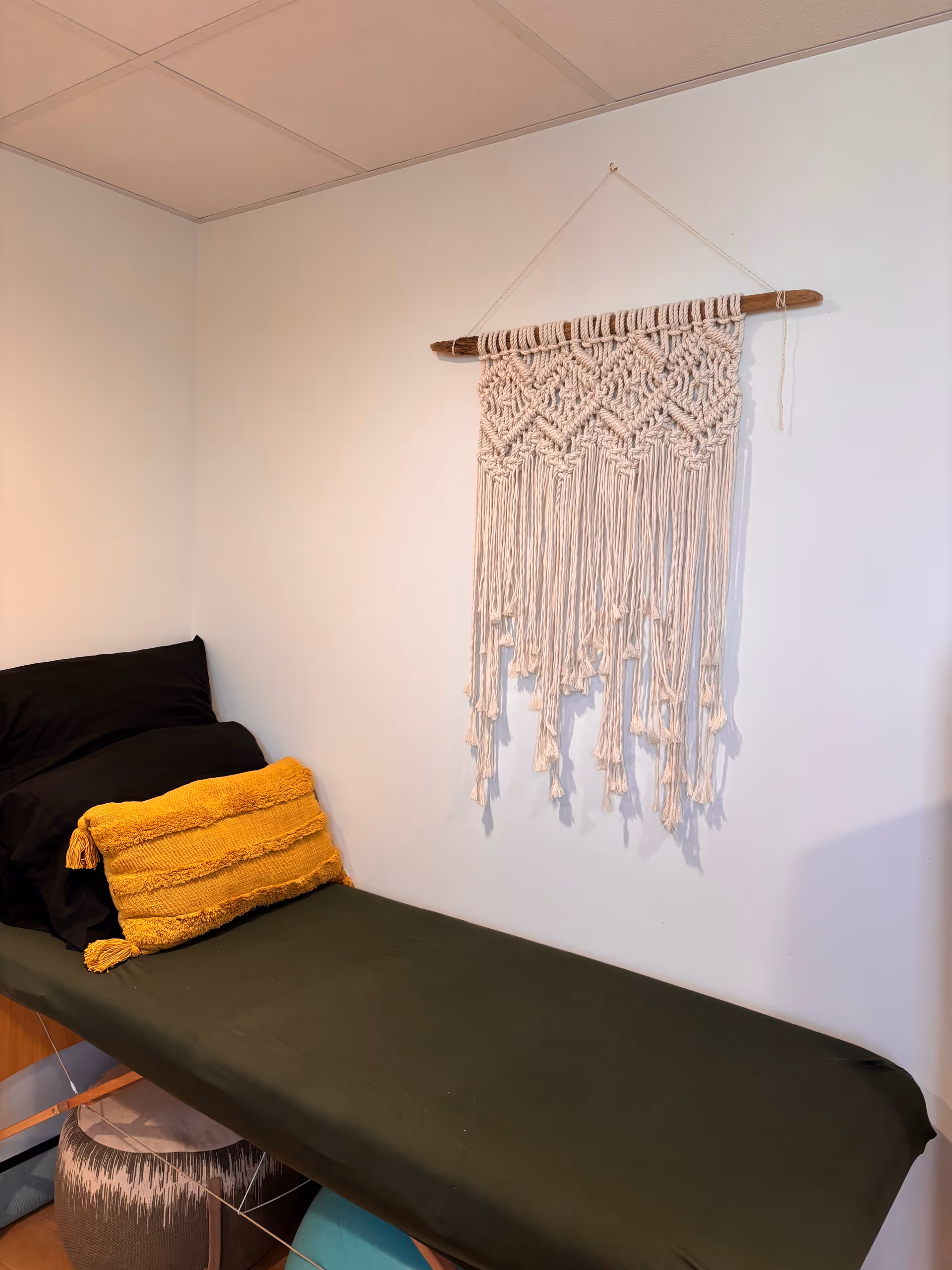 A small tidy room with a single bed, dark green sheet, and macrame wall hanging.