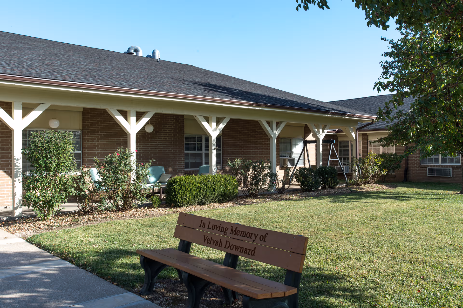 Single-story brick senior living facility with a shaded porch and memorial bench.