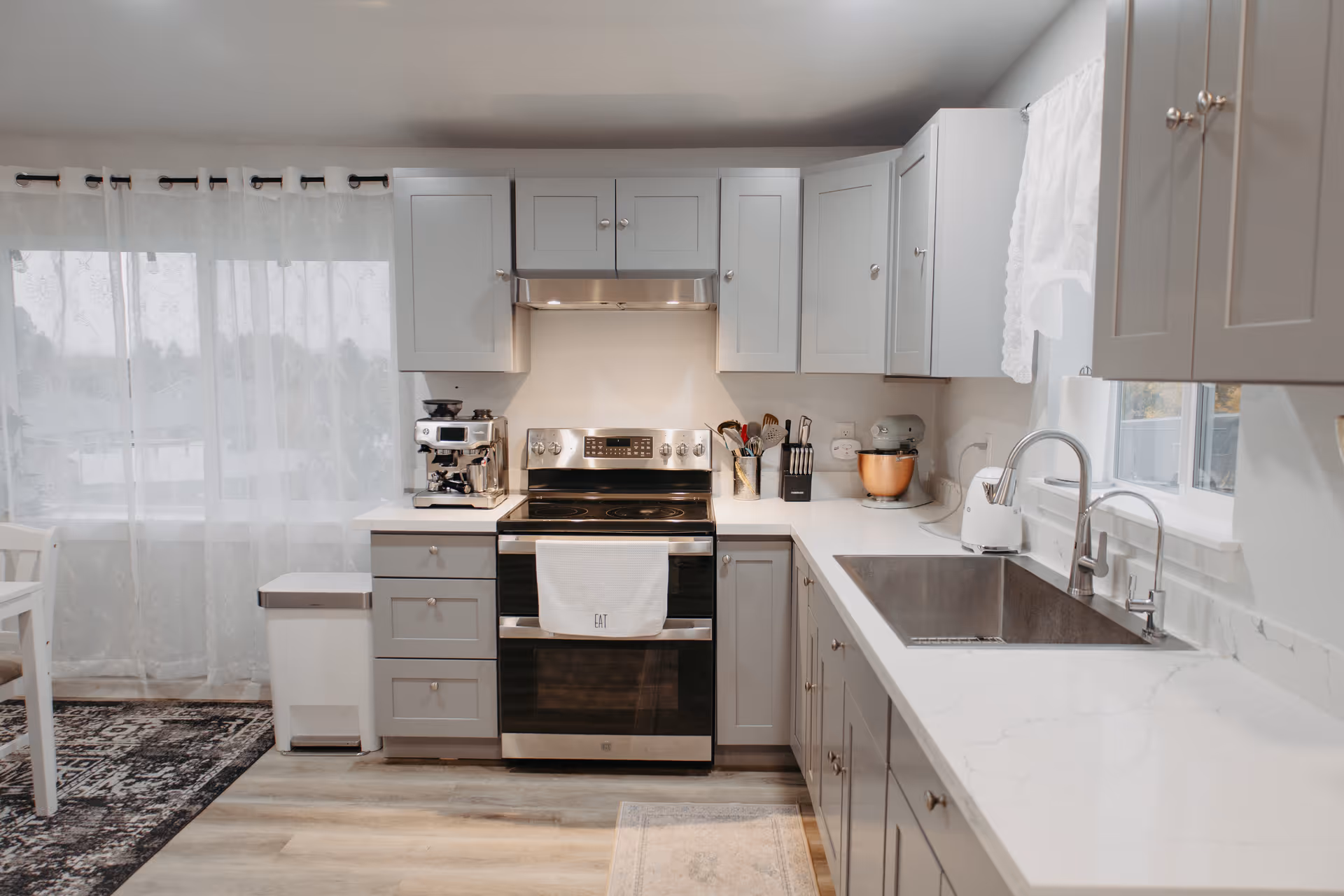 Grey kitchen with stainless steel appliances, white countertops, sink, and sheer-curtained windows.