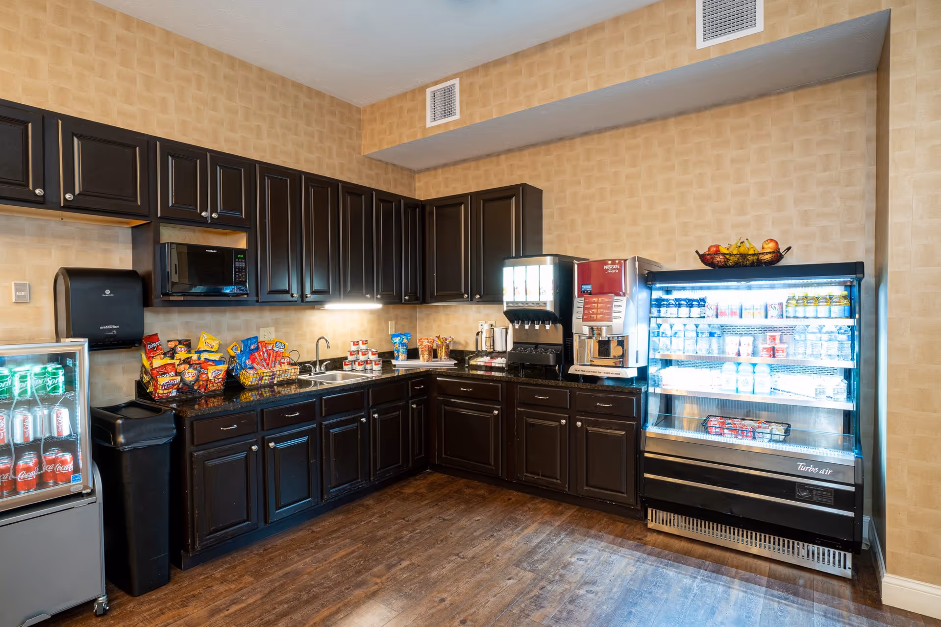 Small kitchenette and snack area with dark cabinets, a refrigerated display, coffee machines and baskets of snacks.
