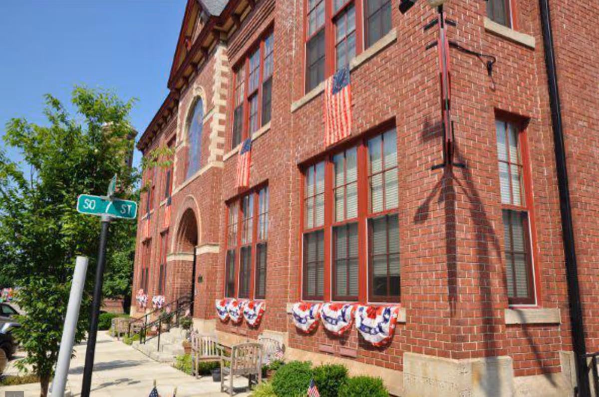 Exterior view of a red brick building decorated with American flags and patriotic bunting. There are benches and small bushes along the sidewalk in front of the building, and a street sign indicating the intersection of South 7th Street and another street.