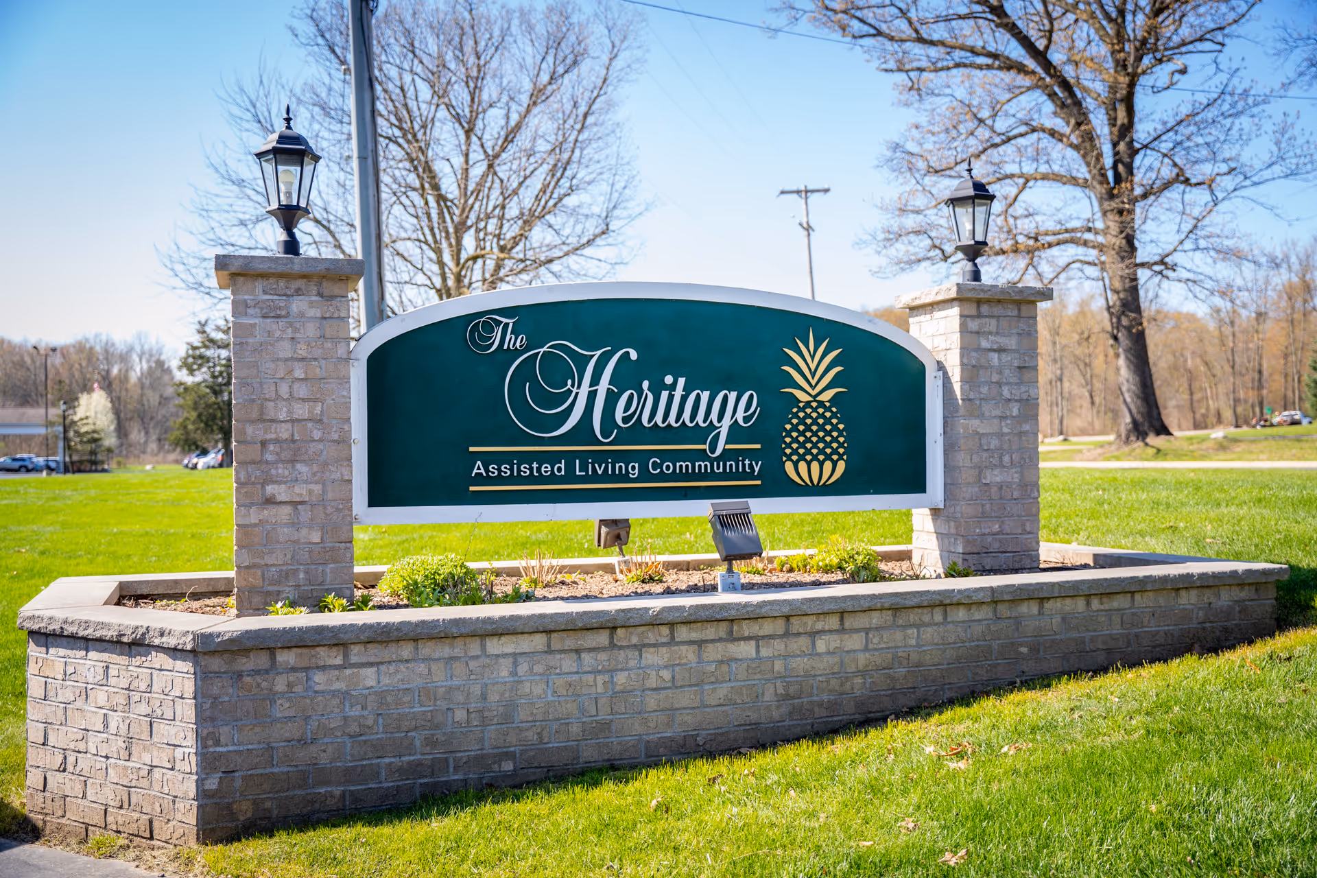 A large green, arched sign reading The Heritage Assisted Living Community sits on a brick pedestal.