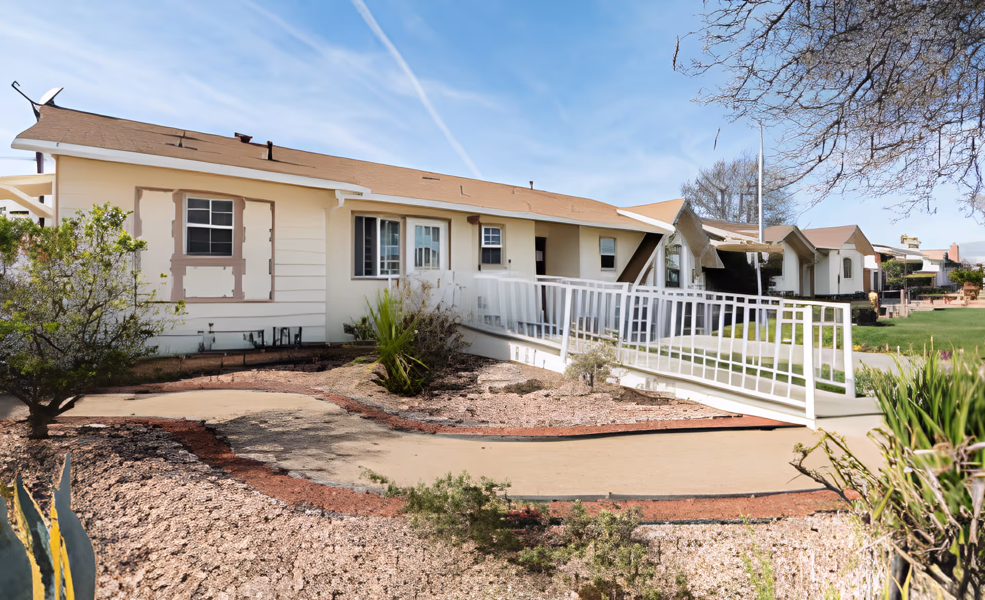 Single-story senior living building with a white accessibility ramp, brown roof, and desert landscaping under a clear sky.
