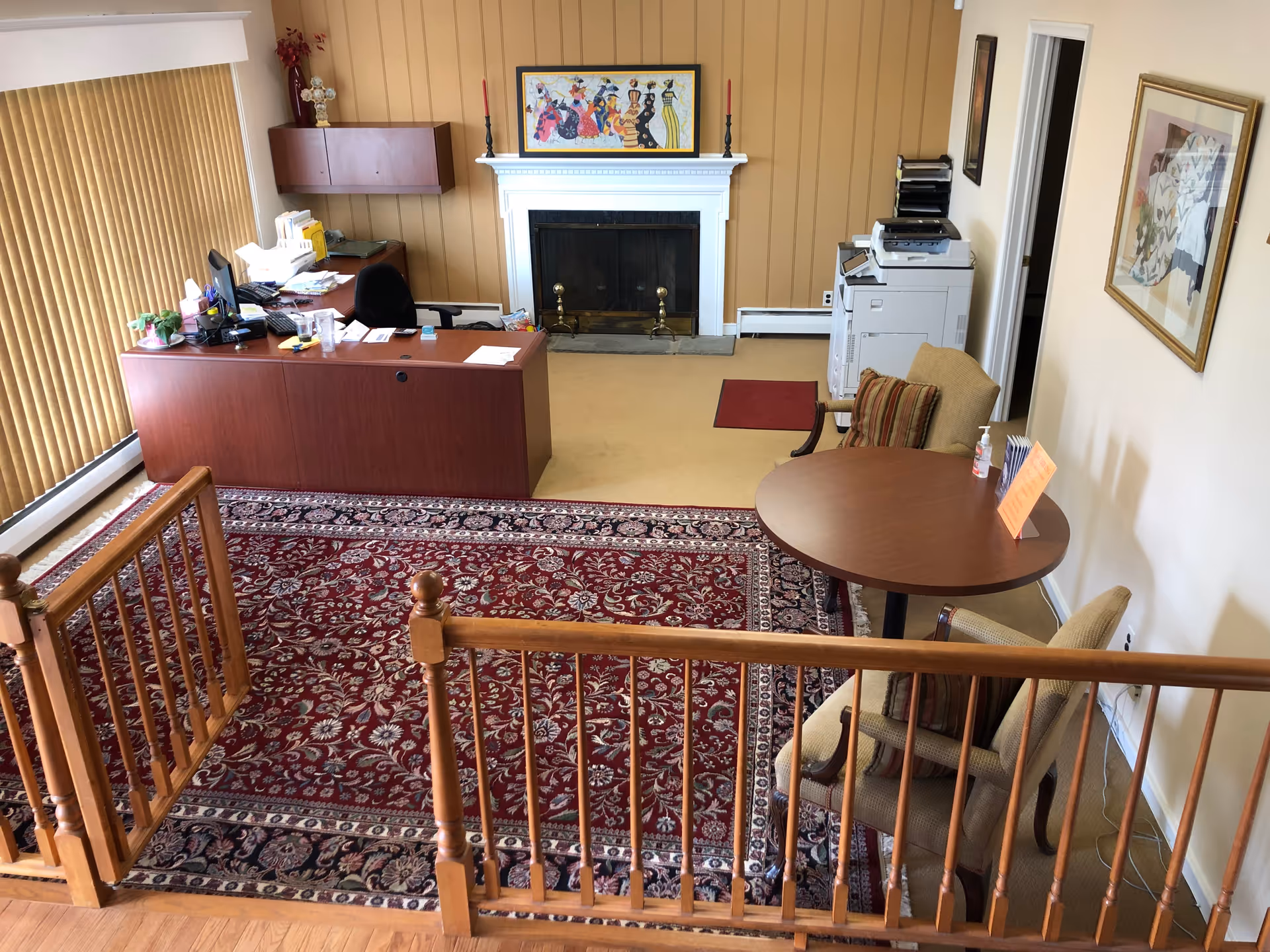 Reception area with wooden desk, fireplace, chairs, round table, and staircase railing.