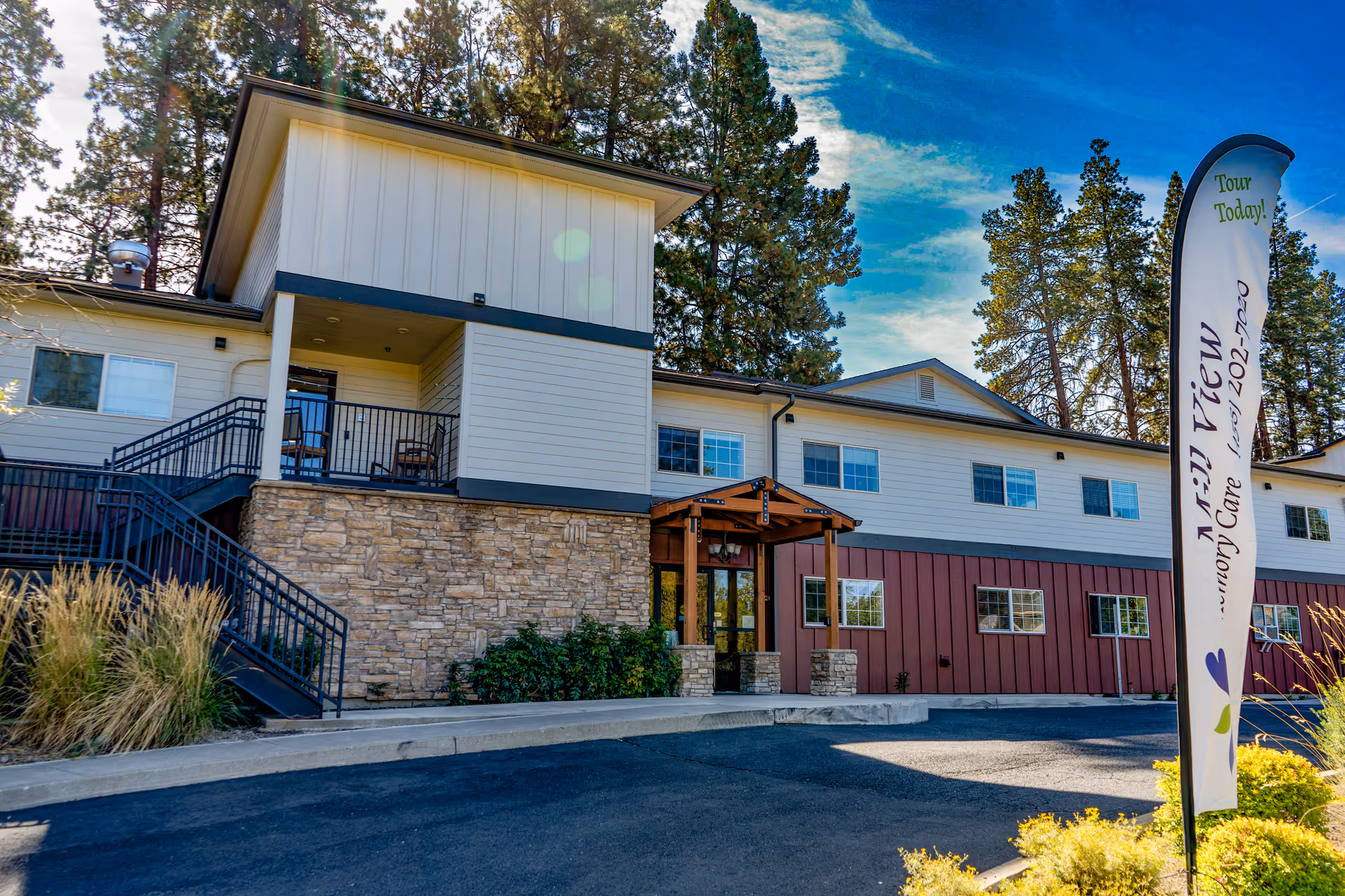 Exterior view of Mill View Memory Care facility showing a two-story building with a stone and siding facade, outdoor stairs, and a covered entrance. There are tall pine trees in the background and a flag sign in the foreground advertising tours today with a phone number.